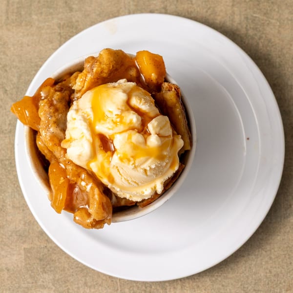 IDA MAE’S peach cobbler dessert with vanilla ice cream and caramel drizzle in a white bowl.